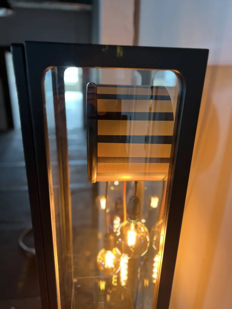 Senta von vorn: schmale Standuhr-Vitrine mit gestreiftem Schirm und Glühbirnen dahinter