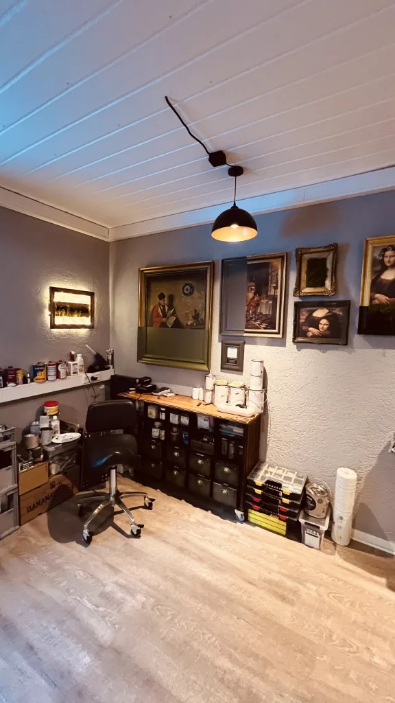 Arbeitsplatz in der Werkstatt: Werkbank mit Farbdosen, Bürostuhl und einer Wand voller gerahmter Bilder